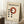 May include: Framed Bauhaus poster with cream background featuring red circle and black dots in geometric arrangement, displayed leaning against white wall corner on textured rug. Coffee cup on wooden block visible nearby, natural lighting creates soft shadows across the minimalist design celebrating the iconic German art school's form follows function philosophy.