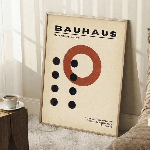 May include: Framed Bauhaus poster with cream background featuring red circle and black dots in geometric arrangement, displayed leaning against white wall corner on textured rug. Coffee cup on wooden block visible nearby, natural lighting creates soft shadows across the minimalist design celebrating the iconic German art school's form follows function philosophy.
