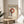 May include: Framed Bauhaus poster with cream background featuring red circle and black dots in geometric arrangement. Wood-framed print displayed on round wooden side table next to beige upholstered chair. Natural sunlight creates shadows across white wall. Green plant leaves visible in corner, creating modern minimalist room styling.