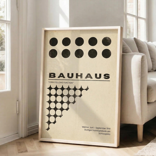 May include: Framed Bauhaus poster with cream background featuring black geometric circles arranged in rows and a diagonal pattern of white circles. The minimalist design displays "Form Follows Function" motto and German text referencing Weimar 1919. Light wooden frame leans against white wall beside beige sectional sofa in bright, airy living room with large windows and natural lighting.