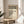 May include: Framed Bauhaus poster with cream background and black geometric circles arranged in rows, featuring "Form Follows Function" text and triangular dot pattern, displayed on light wood round table beside beige upholstered chair with plant leaves visible in corner of minimalist room with white walls and natural lighting.