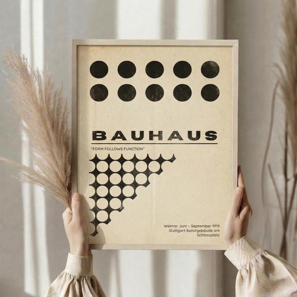 May include: Person in beige clothing holding framed Bauhaus poster with black geometric circles and triangular pattern on cream background, displayed against white curtains with dried pampas grass visible on left side. The minimalist design features bold typography and dots arranged in rows, creating a vintage modernist aesthetic in neutral tones.