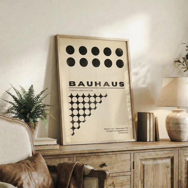 May include: Framed Bauhaus poster with black geometric circles and "Form Follows Function" text displayed on rustic wooden sideboard in neutral-toned living room. Cream upholstered chair, potted fern, ceramic vase, and stacked books create warm, minimalist styling around the vintage-inspired design print in natural wood frame.