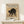 May include: Framed vintage-style poster featuring a black cat in an arched, hissing pose with yellow eyes against aged beige paper background. The print shows Japanese-inspired artwork with traditional calligraphy characters and red seal stamp. Displayed in light wooden frame on white shelf next to small wrapped gift box with twine and dried grass accent. Clean white wall background creates minimalist gallery wall aesthetic.