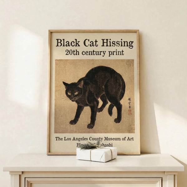 May include: Framed vintage-style poster featuring a black cat in an arched, hissing pose with yellow eyes against aged beige paper background. The print shows Japanese-inspired artwork with traditional calligraphy characters and red seal stamp. Displayed in light wooden frame on white shelf next to small wrapped gift box with twine and dried grass accent. Clean white wall background creates minimalist gallery wall aesthetic.
