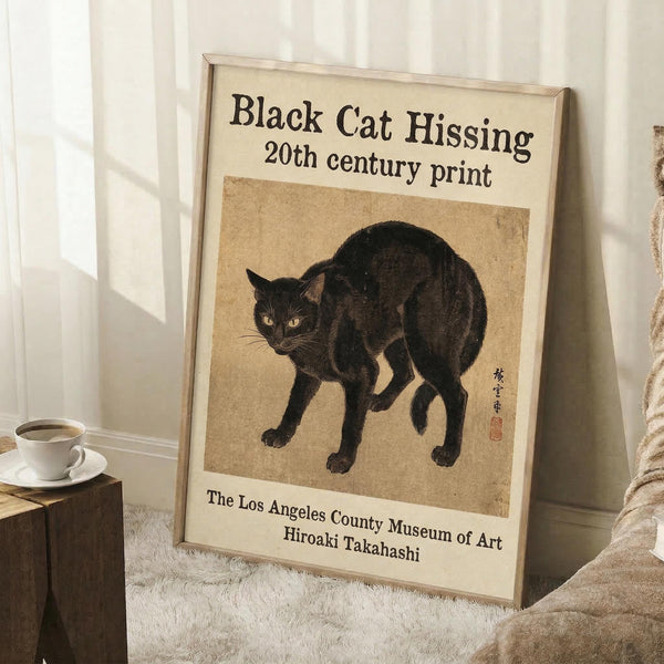 May include: Framed vintage-style poster featuring a black cat with arched back and hissing expression, displayed against white paneled wall. The artwork shows Japanese-inspired illustration on aged beige background with museum attribution text below. Scene includes wooden side table with white coffee cup, textured white rug, and beige knit throw blanket for scale reference.