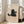May include: Framed poster print featuring a black cat holding a gray mug with marshmallows, illustrated in simple line art style with "Meow" text below, displayed in light wood frame leaning against white wall next to beige upholstered chair in bright, sunlit room with large windows.