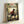 May include: Framed anime poster leaning against white wall showing samurai character in red robe wielding sword in bamboo forest setting. Light wooden frame surrounds colorful artwork with Japanese text and English subtitle about clash of styles and journey. Warm lighting creates shadows on wall behind vintage-style movie poster print.