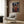 May include: Framed anime poster featuring a dark-haired samurai character in red traditional clothing holding a sword, set against a nighttime street scene with lanterns and crowds. The poster hangs on a wood-paneled wall above a cream upholstered armchair with geometric patterned pillow. Modern brass floor lamp and dark round side table with open book complete the contemporary living room setting.