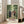 May include: Framed anime poster leaning against white wall next to beige sofa in bright room with large windows. Shows three characters walking through green bamboo forest - man in dark blue traditional robes, woman in pink kimono, and man with afro in red jacket carrying sword. Japanese and English text at bottom. Light wood frame, approximately 2-3 feet tall.