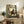 May include: Framed anime poster featuring a dark-haired character in red and black clothing sitting in a bamboo forest setting. The poster displays Japanese text and "A clash of styles. A journey with no map" tagline. Shown leaning against a rustic wooden sideboard alongside books, a ceramic vase, and potted fern in a cozy living room with beige chair and warm lighting.