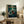 May include: Framed anime poster displayed on rustic wooden cabinet in cozy living room setting. Shows character with flowing red clothing wielding sword in bamboo forest scene. Green and teal artwork with Japanese and English text. Natural wood frame matches cabinet finish. Fern plant, ceramic vase, books, and beige armchair with leather pillows complete the warm, earthy interior decor.