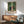 May include: Framed anime poster showing three characters walking through a bamboo forest - a man in dark blue robes, a young woman in pink kimono, and a man with afro in red jacket. Displayed above a wooden sideboard with white ceramic lamp in a modern living room setting with mint green upholstered chair. Warm sunlight streams across white walls creating geometric shadows. Japanese text visible at bottom of artwork.