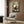 May include: Framed anime poster featuring a dark-haired character in red and black clothing crouched in a bamboo forest setting, displayed on wood-paneled wall above beige upholstered chair with geometric patterned pillow, next to modern brass floor lamp and dark round side table with open book in contemporary living room with natural lighting