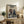 May include: Framed anime poster featuring two samurai warriors with swords positioned over stylized ocean waves in blue and white, with Japanese text at top. Wooden frame sits on rustic wooden cabinet alongside fern plant, books, ceramic vase, and brown leather chair in warm-toned living room setting.
