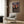 May include: Framed anime poster on wood-paneled wall showing samurai character with sword at harbor dock during sunset. Poster features Japanese text and dramatic purple-orange sky. Modern beige armchair with geometric patterned pillow sits below. Gold floor lamp and dark round side table with open book complete the contemporary living room setting.