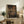 May include: Framed anime poster displaying three characters sitting at a wooden table in a traditional Japanese tea house setting, positioned on a rustic wooden sideboard alongside decorative pottery, books, and a fern plant in a cozy living room with beige armchair and neutral walls