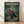 May include: Framed anime poster displayed on light wooden floor against white wall with natural lighting and shadows. Features three samurai characters in dynamic action poses within bamboo forest setting - one in red clothing wielding sword, woman in traditional kimono, man in dark blue robes with glasses. Green forest background with tall bamboo trees, anime art style with bold colors and detailed character designs in natural wood frame.