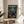 May include: Framed anime poster displayed on round wooden table next to beige upholstered chair. Forest green artwork shows three samurai characters in traditional robes wielding swords in bamboo forest setting. Natural lighting creates shadows across white walls and plant leaves visible in corner.