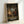 May include: Framed anime poster leaning against white wall showing character in red clothing sitting in traditional Japanese room with tatami mats, wooden architecture, and calligraphy scrolls. Natural wood frame, warm interior lighting, detailed illustration style with earth tones and atmospheric perspective through window opening.