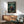 May include: Framed anime-style poster mounted on white wall above wooden credenza, featuring three samurai characters in dynamic fighting poses against bamboo forest backdrop. Green and earth tone artwork with Japanese text, displayed in modern living room with teal upholstered chair, round coffee table, and white ceramic table lamp. Warm natural lighting creates shadows across mid-century modern furniture.