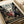 May include: Anime poster print featuring three samurai characters in traditional Japanese clothing wielding swords on a busy street scene. The artwork shows detailed illustration style with muted earth tones and urban backdrop. Print appears flat against white surface with curved white paper edges visible, suggesting product photography setup.