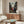 May include: Framed anime poster on white wall above wooden sideboard in modern living room. Poster shows three samurai characters in red and dark clothing on traditional Japanese street. Room features white curved chair, oval glass coffee table, floor lamp, and neutral decor with natural lighting.