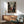 May include: Framed anime poster displayed above wooden sideboard in modern living room setting. Poster shows three characters with swords in traditional Japanese street scene with red and blue clothing against vintage buildings. Mid-century furniture includes teal upholstered chair with wooden arms, round coffee table, white ceramic lamp. Natural sunlight creates shadows on white wall, highlighting the poster's detailed artwork and warm wooden frame.