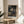 May include: Framed anime poster featuring a long-haired samurai character in blue robes wielding a sword in a traditional Japanese room with tatami mats. Japanese text appears at top and bottom in white characters against the artwork. The wooden-framed print sits on a light wood round side table next to a beige upholstered chair, with a potted plant visible and natural window light creating soft shadows on neutral walls.