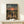 May include: Framed anime poster displayed on light wooden shelf against white wall. Artwork shows three characters walking down dirt road at sunset with rolling hills and orange sky. Small white wrapped gift box sits in front of frame. Warm earth tones throughout vintage-style illustration.