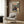 May include: Framed anime poster mounted on wood-paneled wall showing samurai character in red and blue clothing wielding sword in mountainous landscape. Japanese text at top with English subtitle. Cream upholstered armchair with geometric patterned pillow positioned below. Gold floor lamp and dark round side table with open book complete modern living room setting.