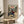 May include: Framed anime poster featuring a black-haired samurai character in red and blue clothing wielding a sword, set against a mountainous landscape with green trees. The artwork displays Japanese text at the top and "The Revolution of a Generation" subtitle. The thin wooden frame rests on a round wooden side table next to a beige upholstered chair, with a plant casting shadows on the white wall behind in natural sunlight.