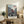 May include: Framed anime poster displayed on rustic wooden console table alongside fern plant, books, and ceramic vase. Artwork shows dynamic samurai character in red clothing wielding sword in traditional Japanese street scene with onlookers. Japanese text at top and bottom. Light wood frame against cream wall in cozy living room setting with beige upholstered furniture and warm lighting.