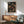 May include: Framed anime poster showing a samurai character with dark hair in red and gray clothing, holding a sword while crouching in a lantern-lit Japanese street scene. The artwork features warm orange lighting and traditional architecture. Displayed above a wooden sideboard with white ceramic lamp, next to a mid-century modern green upholstered chair with wooden arms in a minimalist living room with white walls and natural sunlight streaming across the space.