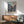 May include: Framed anime poster featuring a samurai character in red clothing wielding swords in an action scene with Japanese text, displayed above a wood sideboard with white ceramic lamp in a modern living room setting with mint green upholstered chair and small round coffee table, soft natural lighting on white walls.