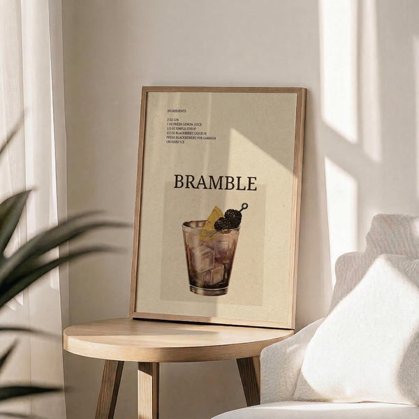 May include: Framed beige poster displaying a bramble cocktail recipe with ingredients list at top and large "BRAMBLE" text above illustrated purple cocktail glass garnished with blackberries and lemon. Wood frame sits on round wooden side table next to cream upholstered chair with throw pillow. Green plant leaves visible in corner with natural sunlight creating soft shadows on white wall.