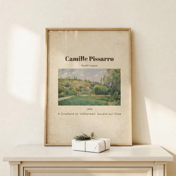 May include: Framed art print featuring Camille Pissarro's 1874 impressionist landscape painting of a pastoral countryside scene with rolling green hills, trees, and a cowherd. The beige poster has black text identifying the artist, museum, date and title. Displayed in a thin wooden frame on a light wood shelf beside a small white wrapped gift with twine and pine sprig against a cream wall.