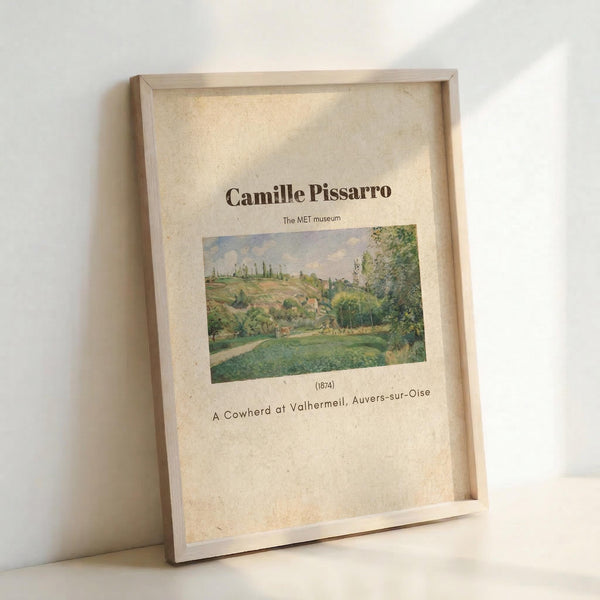 May include: Framed art print leaning against white wall showing Camille Pissarro's pastoral landscape painting from 1874. The artwork depicts rolling green hills, trees, and countryside in soft impressionist colors. Print features museum-style layout with artist name, small landscape image, date, and title on cream textured background. Light wood frame casts gentle shadow on white surface.