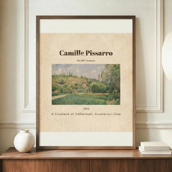 May include: Framed art print featuring Camille Pissarro's 1874 landscape painting of a cowherd in Valhermeil, Auvers-sur-Oise. The poster shows rolling green hills, trees, and pastoral countryside in impressionist style with muted greens and earth tones. Displayed in thin brown frame on wooden sideboard beside white ceramic vase and books against cream paneled wall.