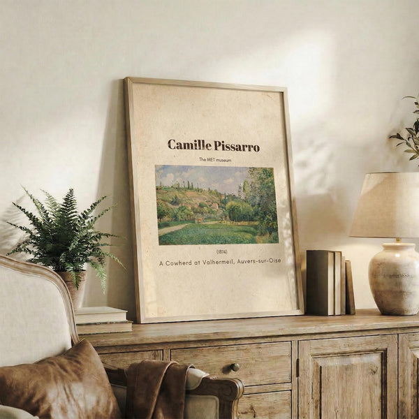 May include: Framed art print of Camille Pissarro's impressionist landscape painting featuring green countryside with rolling hills and trees, displayed on rustic wooden sideboard alongside fern plant, stacked books, and ceramic pottery in neutral-toned living room setting with upholstered chair visible.