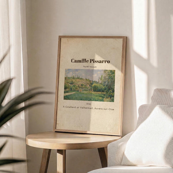 May include: Framed art print featuring Camille Pissarro's 1874 pastoral landscape painting displayed on a light wood round side table next to a cream upholstered chair. The beige poster shows a countryside scene with green fields, trees, and buildings under a cloudy sky. Natural sunlight creates soft shadows across the neutral-toned room setting with white curtains and plant leaves visible in the corner.