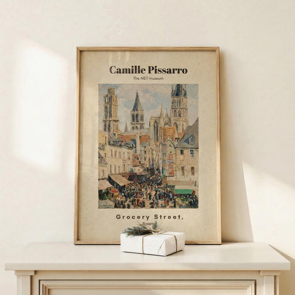 May include: Framed reproduction of Camille Pissarro's impressionist painting showing a bustling French market street scene with Gothic cathedral towers, colorful buildings, and crowds of people. The vintage-style poster features cream matting in a light wood frame, displayed on a pale wooden surface alongside a small white gift box with natural twine and greenery against a neutral wall.