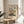 May include: Framed art print displayed on round wooden side table next to beige upholstered chair. Print features Impressionist street scene painting with crowds of people walking through historic European city square surrounded by Gothic cathedral towers and traditional buildings in muted earth tones. Cream matting with brown text in thin gold frame. Soft natural lighting creates shadows across neutral-toned room with plant leaves visible in corner.