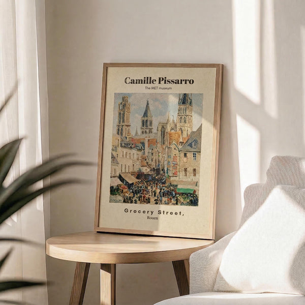 May include: Framed art print displayed on round wooden side table next to beige upholstered chair. Print features Impressionist street scene painting with crowds of people walking through historic European city square surrounded by Gothic cathedral towers and traditional buildings in muted earth tones. Cream matting with brown text in thin gold frame. Soft natural lighting creates shadows across neutral-toned room with plant leaves visible in corner.
