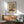 May include: Framed museum poster of Impressionist street scene showing bustling market with Gothic cathedral towers, colorful medieval buildings, and crowds of people in muted blues, oranges and earth tones. Displayed above mid-century modern walnut credenza with ceramic lamp, next to sage green upholstered armchair with wooden frame in bright, naturally-lit room with white walls.