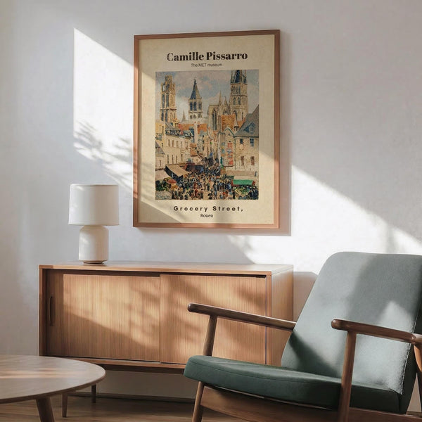 May include: Framed museum poster of Impressionist street scene showing bustling market with Gothic cathedral towers, colorful medieval buildings, and crowds of people in muted blues, oranges and earth tones. Displayed above mid-century modern walnut credenza with ceramic lamp, next to sage green upholstered armchair with wooden frame in bright, naturally-lit room with white walls.