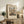 May include: Framed vintage art poster featuring a bustling European street scene with Gothic cathedral towers, colorful medieval buildings, and crowds of people below. The print sits on a rustic wooden console table alongside potted plants, ceramic vases, and stacked books. Warm natural lighting illuminates the neutral-toned living room setting with a cream upholstered chair visible in the corner.