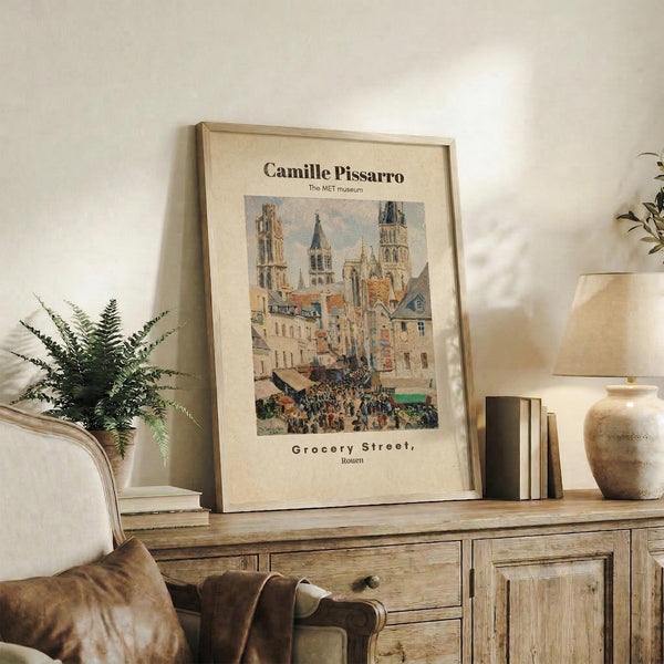 May include: Framed vintage art poster featuring a bustling European street scene with Gothic cathedral towers, colorful medieval buildings, and crowds of people below. The print sits on a rustic wooden console table alongside potted plants, ceramic vases, and stacked books. Warm natural lighting illuminates the neutral-toned living room setting with a cream upholstered chair visible in the corner.