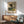 May include: Framed poster of an impressionist landscape painting showing tall trees beside a winding path at sunset, with golden and green tones throughout the pastoral scene. The artwork is displayed above a wooden sideboard with sliding doors, next to a white ceramic table lamp and mid-century modern green upholstered armchair with wooden frame. Natural sunlight creates geometric shadows across the white wall and wooden furniture in this contemporary living room setting.