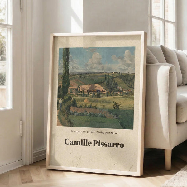 May include: Framed art print of a pastoral countryside scene with rolling green fields, farm buildings, and cloudy blue sky in impressionist style, displayed in light wood frame leaning against white wall next to beige upholstered chair in bright, airy living room with large windows and natural sunlight streaming across hardwood floors.